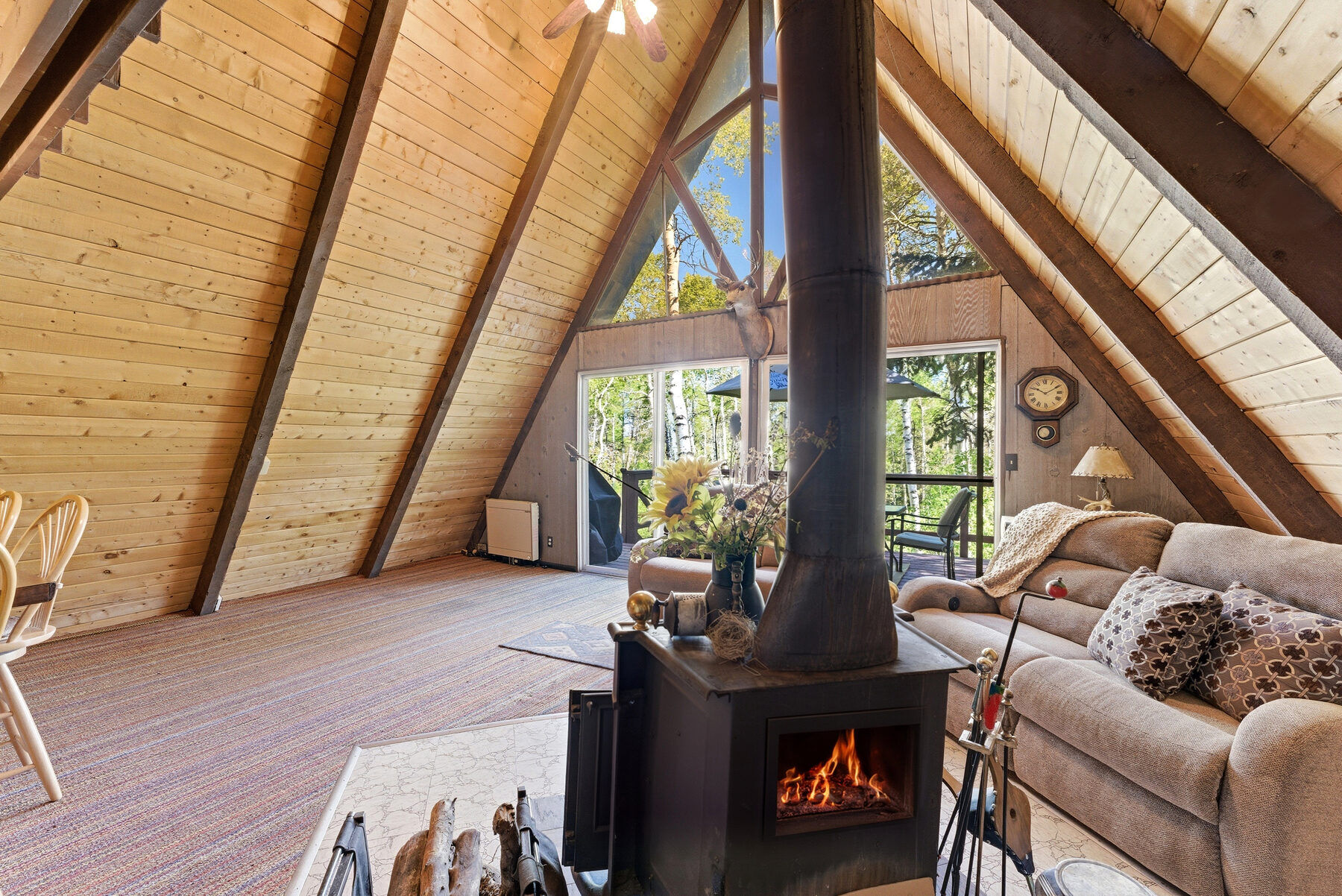 Vaulted A-frame great room with lit wood-burning stove and floor-to-ceiling forest windows